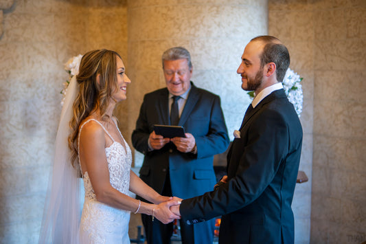 Man and woman looking into each other's eyes, holding hands during their wedding day, with the woman wearing a beautiful white dress and the man in a dark black suit. Their wedding administrator reads their vows in front of a large sandstone brick wall, creating a heartfelt and memorable moment.