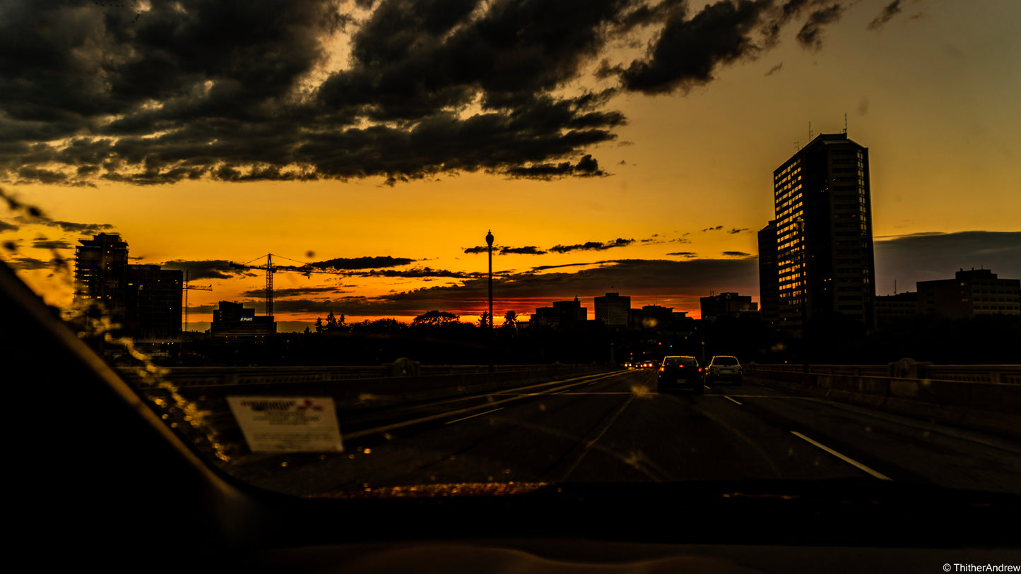 Dark sunset over a city skyline with a silhouette of a car driving over a bridge. The car's windows reflect the glowing light from skyscrapers as seen through the car's side window, capturing the essence of an urban evening.