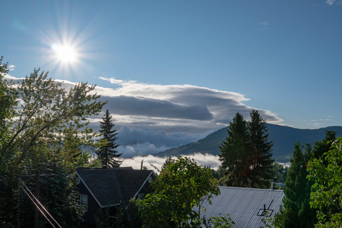 Sun in a blue sky reaching over a dense collection of thick clouds flowing over a bright green mountain town, capturing a contrast between the vivid natural beauty and dramatic sky.