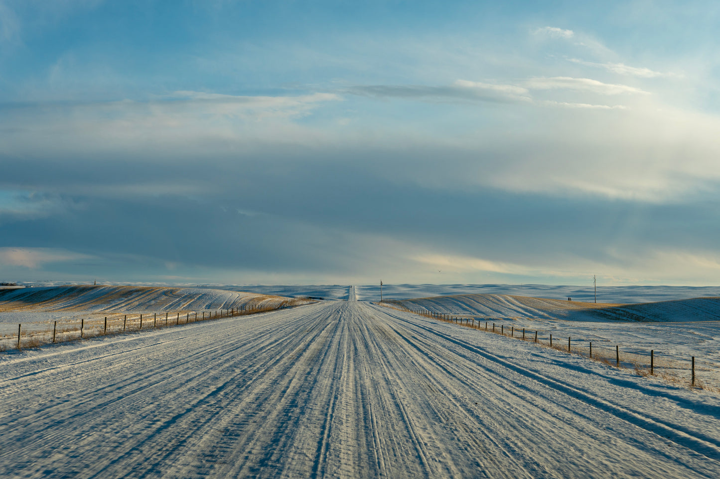 Straight snow-packed gravel road leading through rolling snow-covered prairie fields, creating a peaceful winter landscape with vast open spaces.